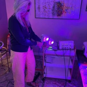 A person with long white hair, wearing sunglasses, a dark cardigan, light yellow pants, and sandals, stands indoors holding a device emitting purple light, near medical equipment and abstract artwork—perhaps studying ticks in summer.