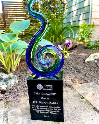 A colorful glass swirl sculpture sits atop a black plaque reading: Global Lyme Alliance Service Award Honoring Dr. Susan Marra…2024, surrounded by green plants in a garden setting.