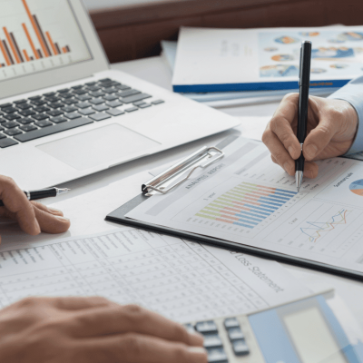Two people work at a desk with financial charts, graphs, and documents. One person holds a pen over a clipboard, while the other uses a calculator. A laptop and more papers with data are also visible.