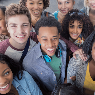 A diverse group of young adults, smiling and standing close together, look up at the camera. One person in front wears blue headphones around his neck. The group appears friendly and cheerful.