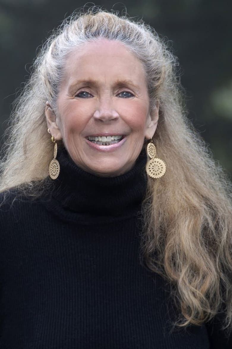 An older woman with long, wavy blonde hair smiles at the camera. She is wearing a black turtleneck sweater and large, intricate gold earrings. The background is outdoors and softly blurred.
