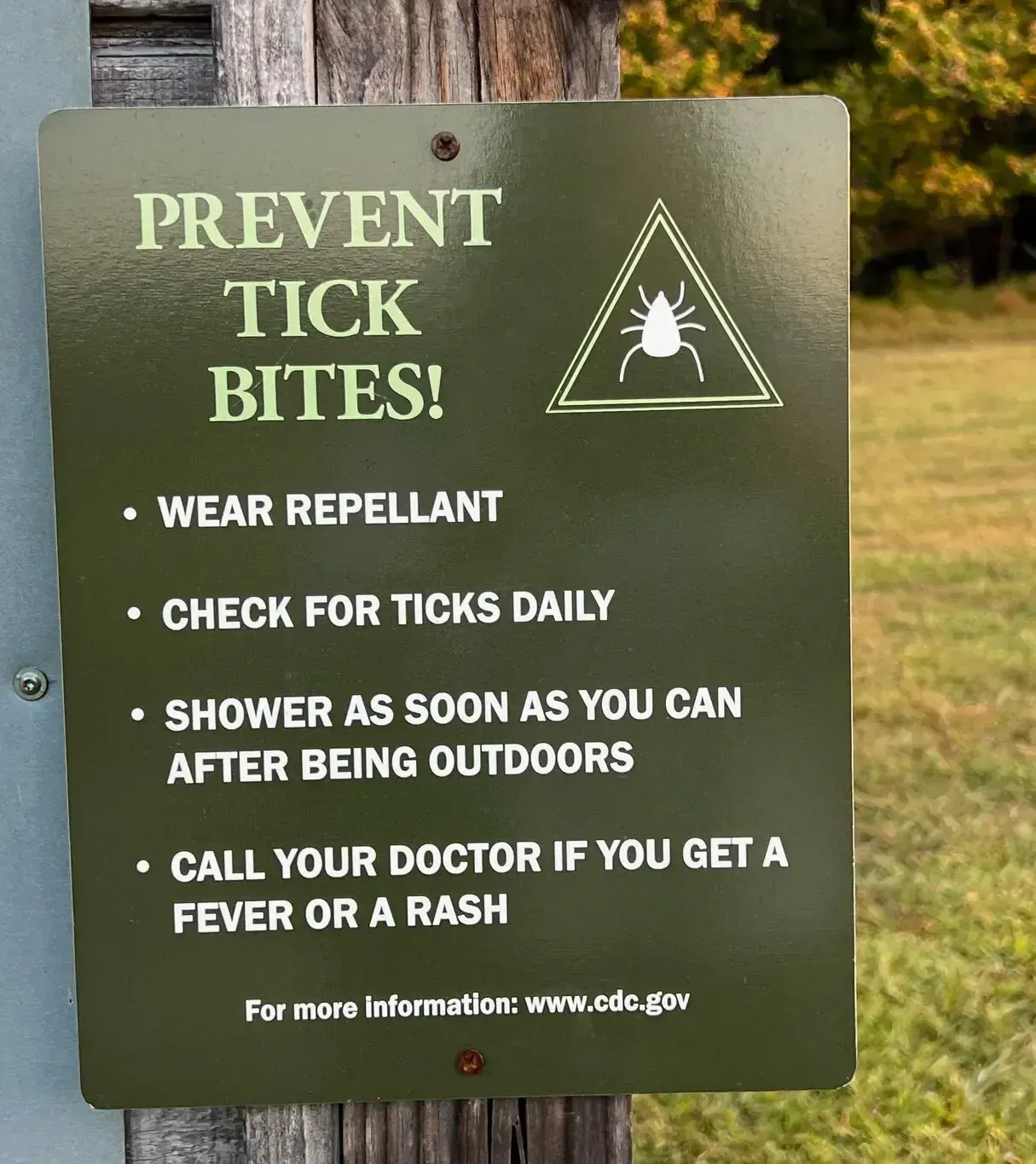 Green sign on a wooden post reads Prevent tick bites! with tips: wear repellent, check for ticks, shower after being outdoors, and call your doctor if you get a fever or rash. A field and bench are in the background.