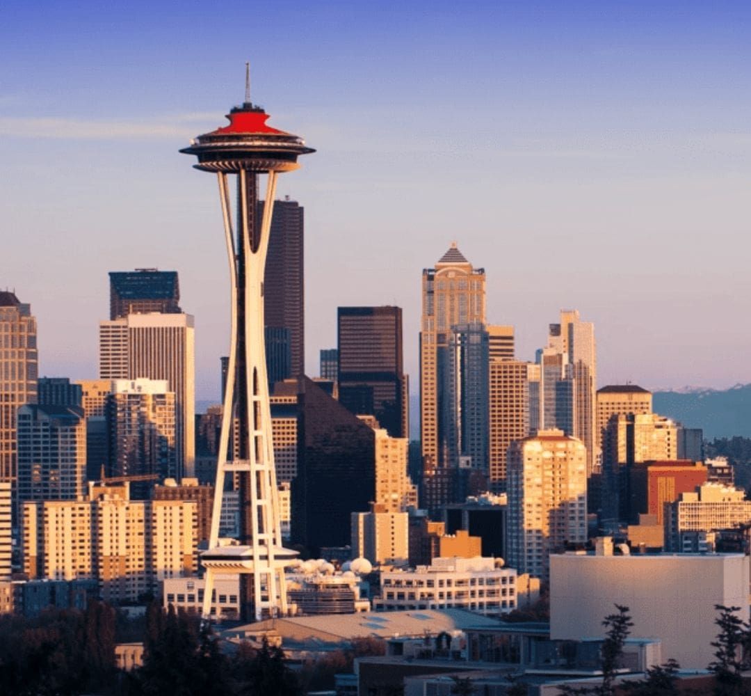 Seattle skyline at sunset featuring the Space Needle in the foreground, surrounded by modern skyscrapers and Mount Rainier visible in the distant background under a clear sky.