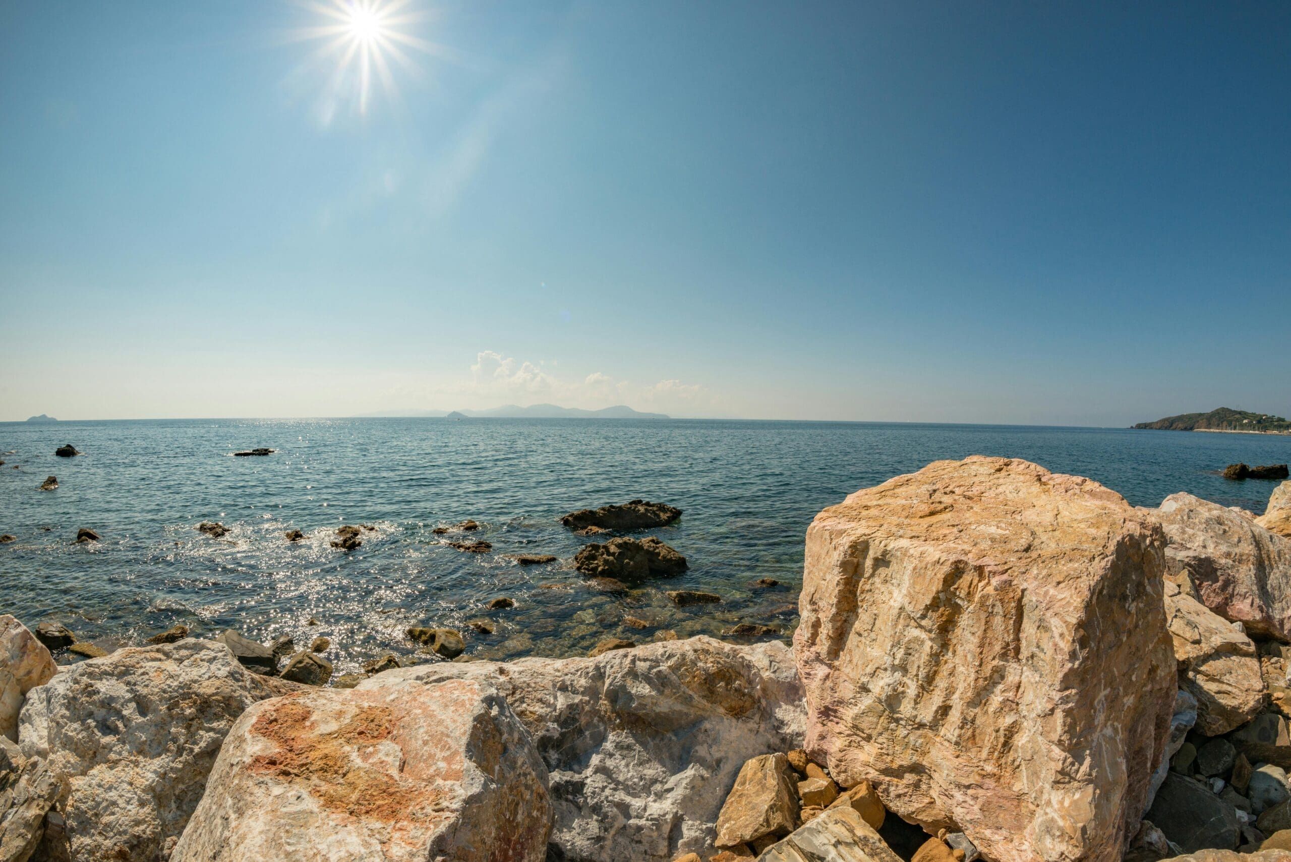Large sunlit rocks in the foreground by a clear, blue sea with scattered smaller rocks in the water, under a bright sky with the sun shining and distant land visible on the horizon.