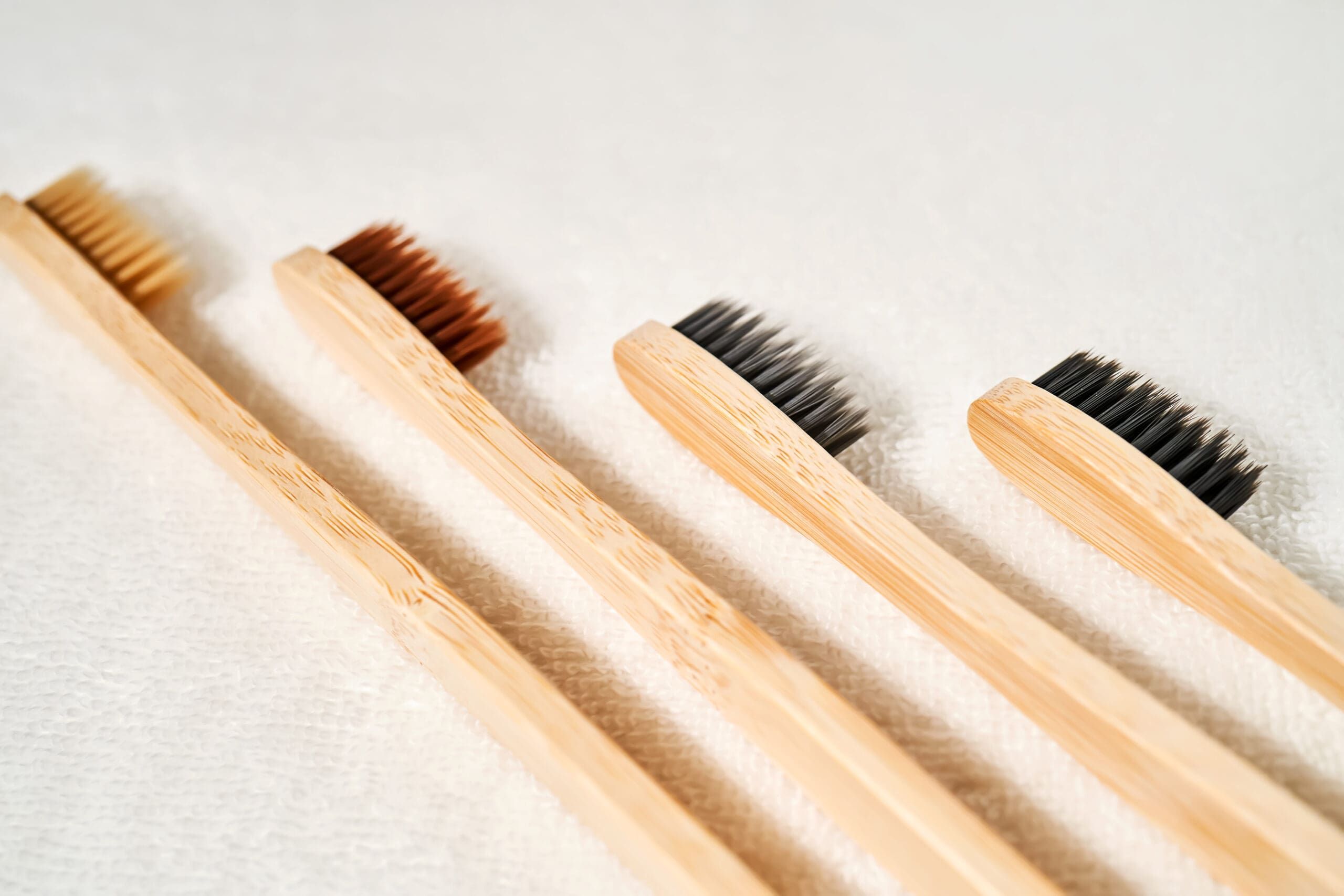 Four bamboo toothbrushes with different colored bristles (brown, dark brown, gray, and black) are lined up diagonally on a white textured surface.