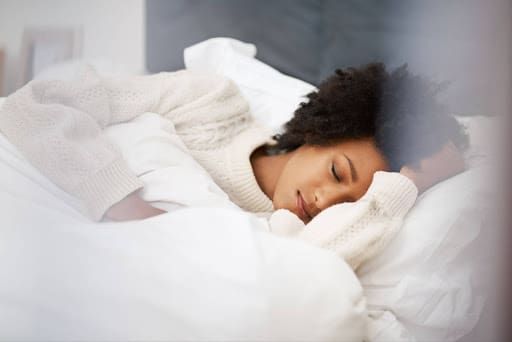 A woman with curly hair is peacefully sleeping on her side in bed, wearing a cozy white sweater and a Neuroradiant 1070 Red Light Helmet, covered with a soft white blanket.