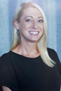 A woman with straight, long blonde hair wearing a black short-sleeve top smiles at the camera. She has blue eyes and is standing in front of a light blue curtain background.our office manager