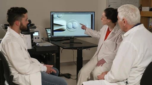 Three scientists in lab coats sit around a screen. One person points at an image of cells related to red light therapy for Lyme disease on the monitor, while the others watch attentively. Lab equipment and notes are visible in the background.