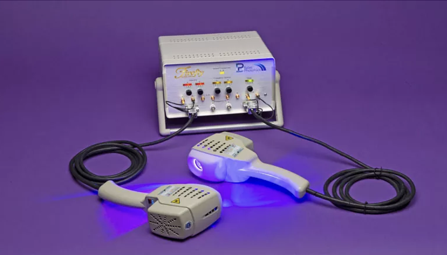 A medical device with a white control unit and two handheld applicators connected by cables, all placed on a purple surface with a purple background. The device emits blue light from the applicators.