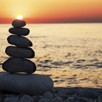 A stack of smooth stones balanced on a rocky beach with the sun setting over calm ocean waves in the background, creating an orange and golden sky.