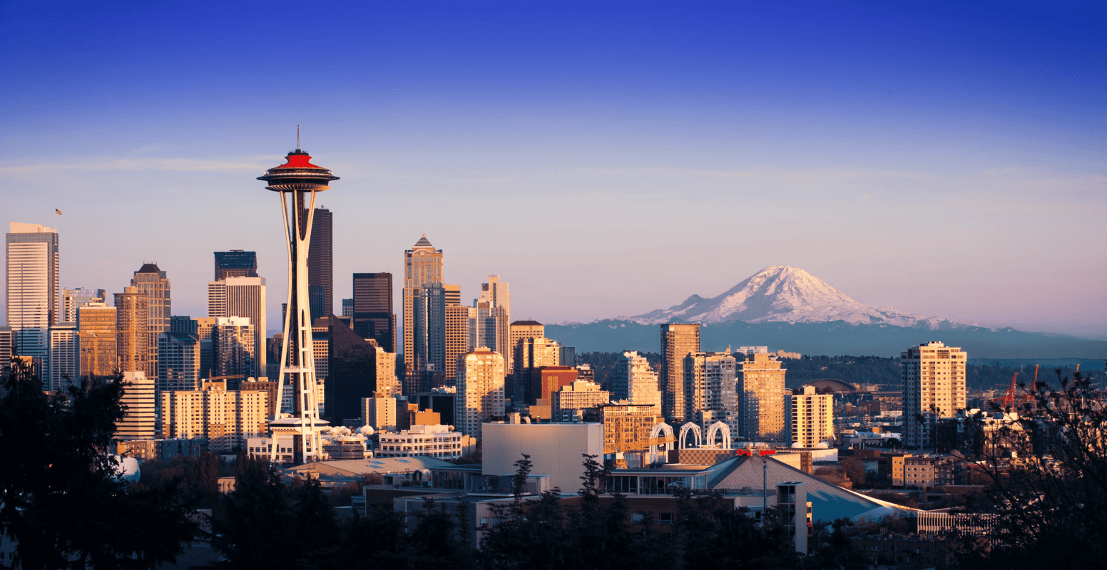 Seattle skyline at sunset with the Space Needle in the foreground, tall buildings, and Mount Rainier covered in snow visible in the background under a clear, blue sky.