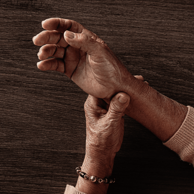 Close-up of an elderly persons hands, one holding the others wrist, against a wooden surface. The person wears a bracelet and a long-sleeve sweater, suggesting care, pain, or tenderness. parkinsons disease