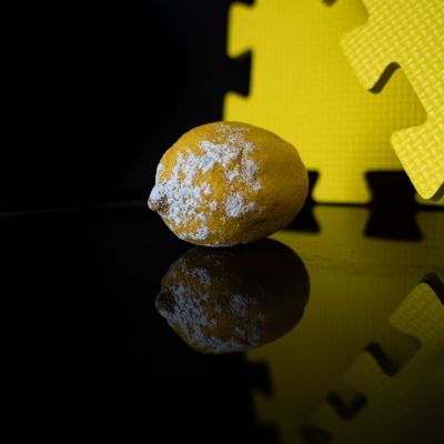 A moldy lemon with white patches sits on a reflective black surface next to yellow interlocking foam mats against a dark background.