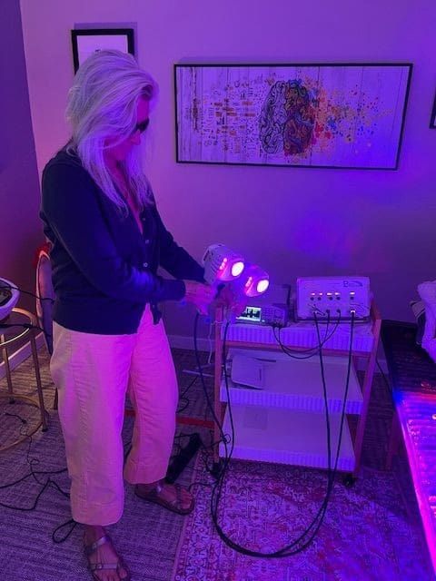 A person with long white hair, wearing sunglasses, a dark cardigan, light yellow pants, and sandals, stands indoors holding a device emitting purple light, near medical equipment and abstract artwork—perhaps studying ticks in summer.