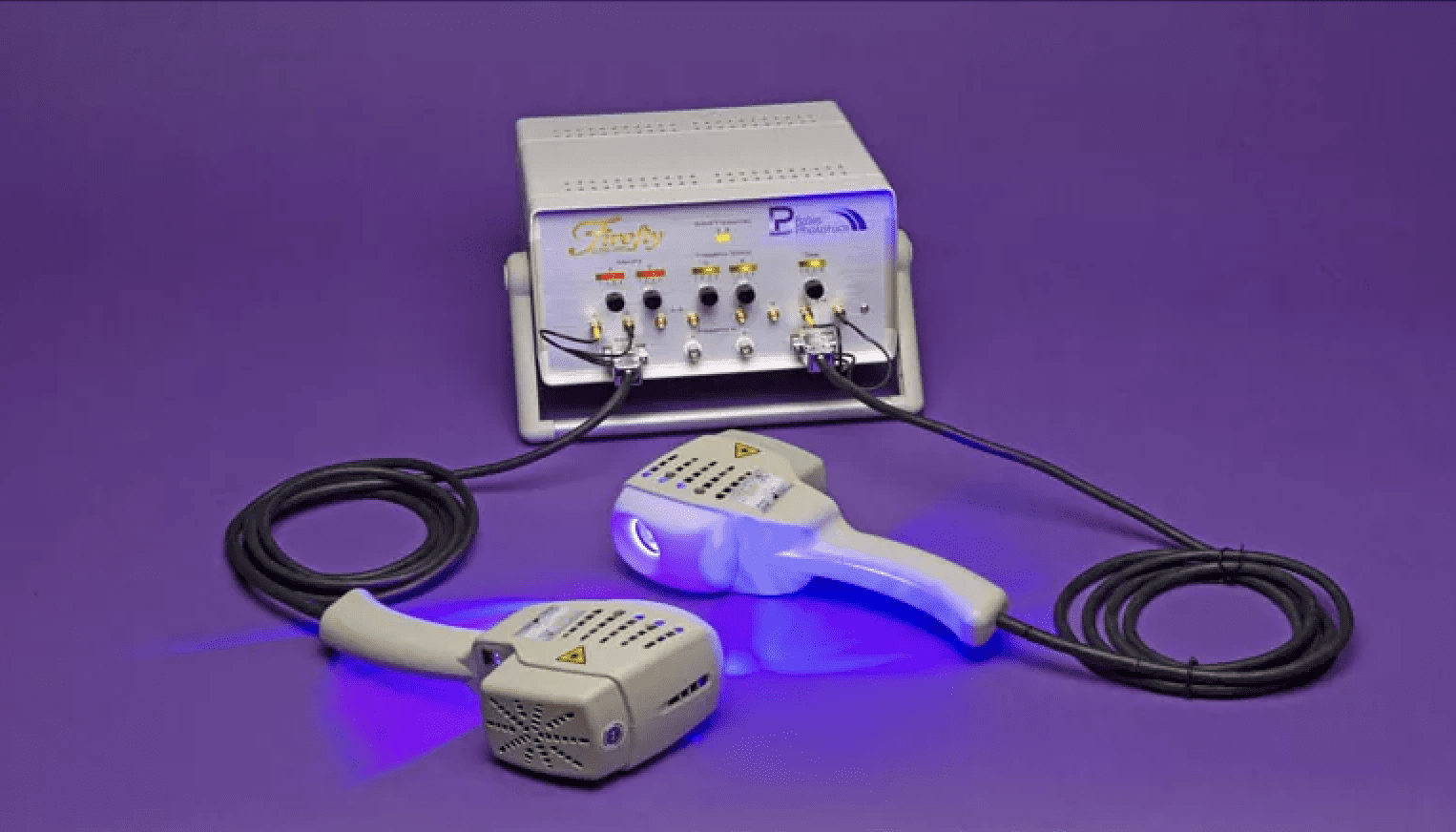 A medical device with a white control unit and two handheld applicators connected by cables, all placed on a purple surface with a purple background. The device emits blue light from the applicators.
