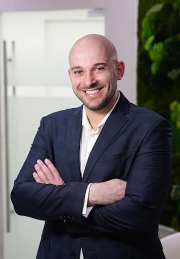 A bald man in a dark suit and white shirt stands indoors with arms crossed, smiling confidently at the camera. The background features a glass door and a wall with green plants.