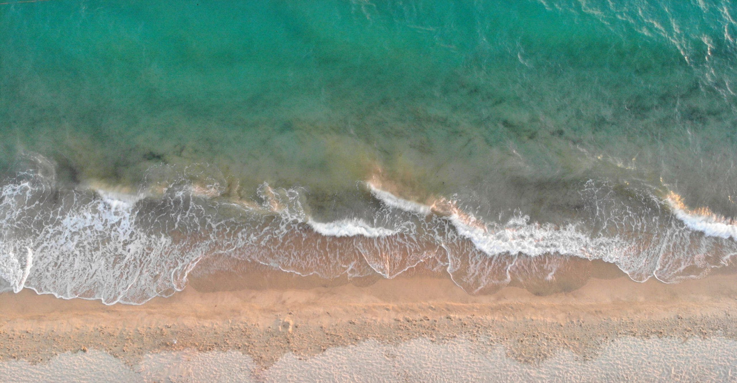 Aerial view of gentle waves crashing on a sandy beach with turquoise water and foamy white surf along the shoreline.