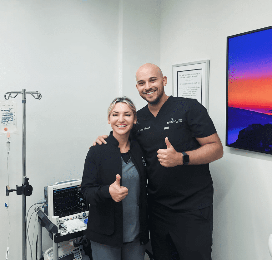 Two healthcare professionals in scrubs smile and give thumbs up in a medical room with monitors, IV pole, and medical equipment. A framed certificate and a colorful landscape photo are on the walls.