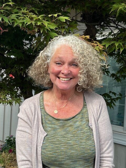 A smiling woman with curly gray hair stands outdoors in front of leafy green plants, wearing a green top, a light cardigan, and a gold necklace.