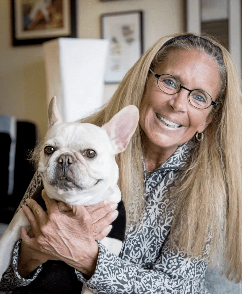 Dr Susan Marra, a lyme literate doctor in Seattle, holds a white French Bulldog on her lap. The woman wears a patterned jacket and sits indoors with framed pictures in the background.