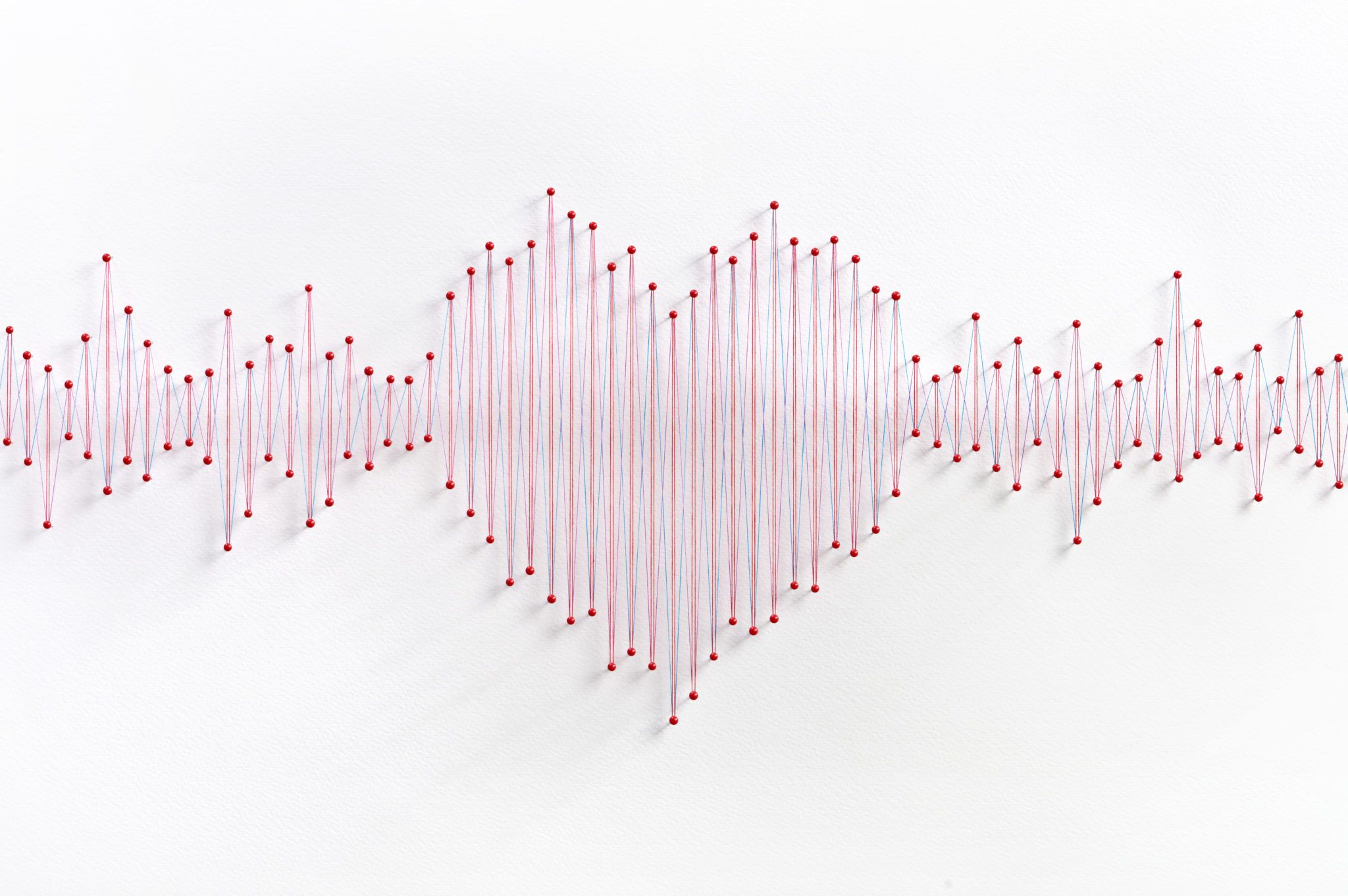 Red string forms a heart shape in the center of a pulsating heartbeat line, created by string stretched between red pins on a white background.