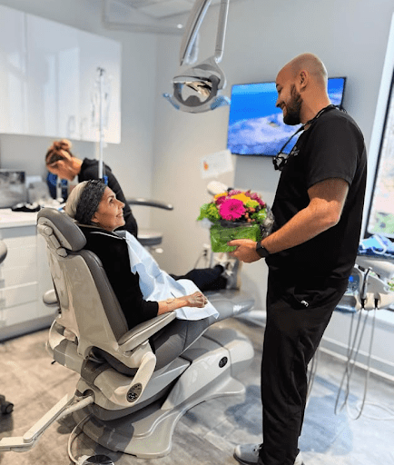 A woman sits in a dental chair smiling at a man holding a bouquet of flowers, while another person works at a desk in the background in a bright, modern dental clinic.