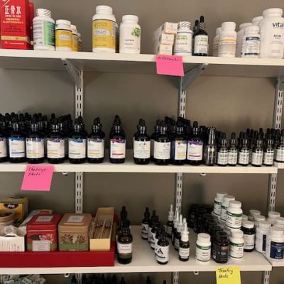 Shelves displaying various health supplements, bottles, and boxes. Several items have handwritten pink and yellow sticky notes labeling categories like antivirals, challenge herbs, and tonify pack.