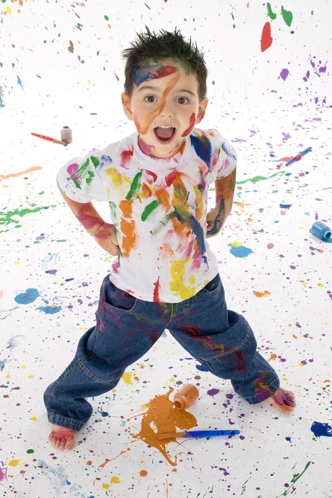 A young boy stands barefoot on a white floor covered in colorful paint splatters, wearing jeans and a paint-stained white shirt, with paint on his face and hands, smiling and looking excited.