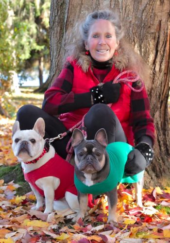 A smiling woman in a red plaid vest sits by a tree on autumn leaves, holding two French Bulldogs—one wearing a red sweater, the other a green one. contact us