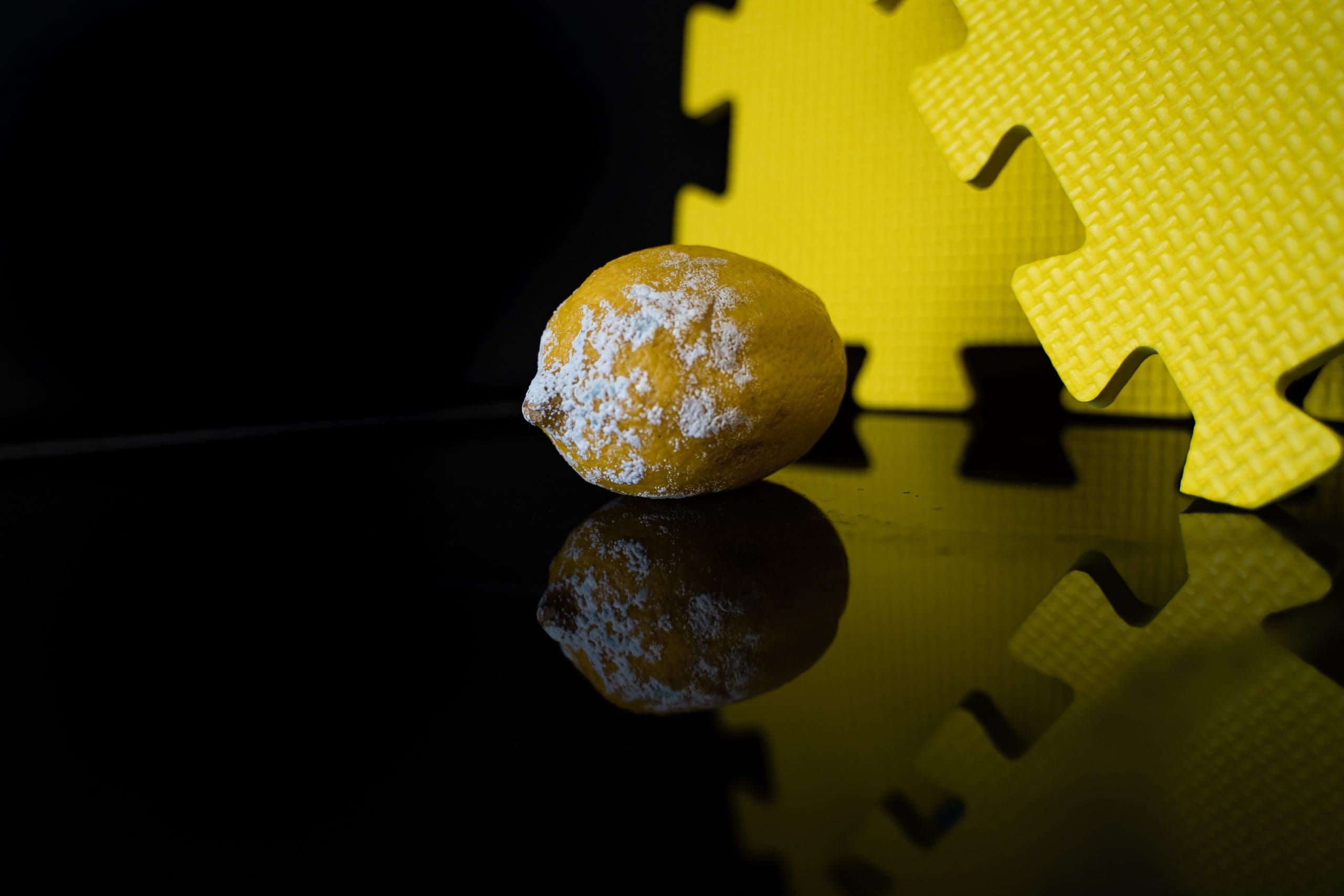 A moldy lemon with white patches sits on a reflective black surface next to yellow interlocking foam mats against a dark background.