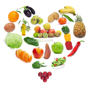 A variety of fruits and vegetables arranged in the shape of a heart, symbolizing healthy eating. Items include bananas, apples, lettuce, peppers, pineapple, tomatoes, kiwi, and more, with a salad bowl at the bottom right.