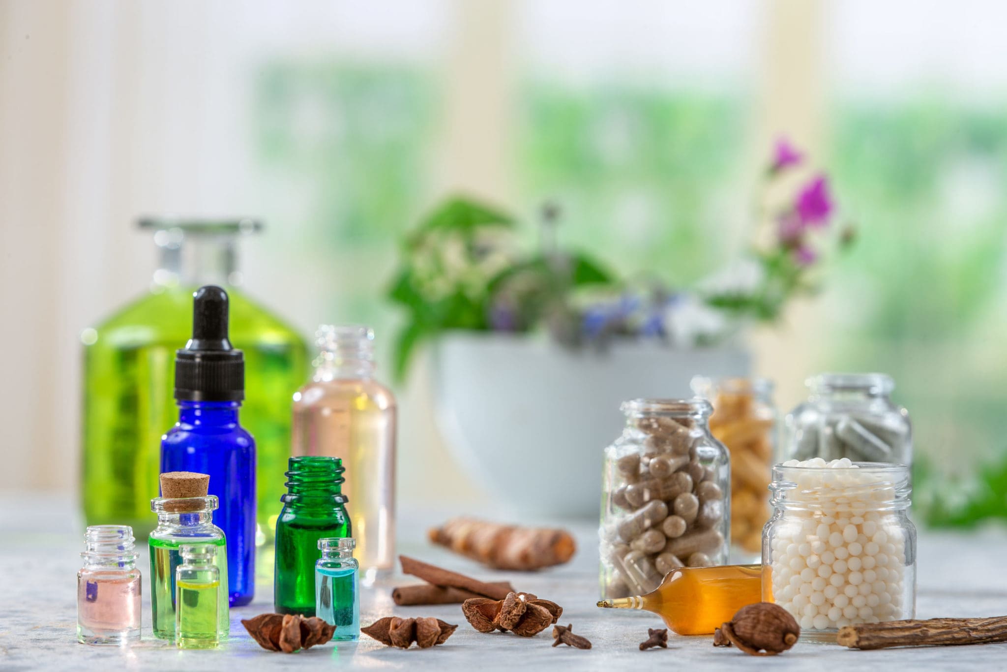 An assortment of small bottles with colored liquids, jars of capsules and tablets, and natural ingredients like cinnamon and star anise on a table, with a blurred bowl of flowers in the background.