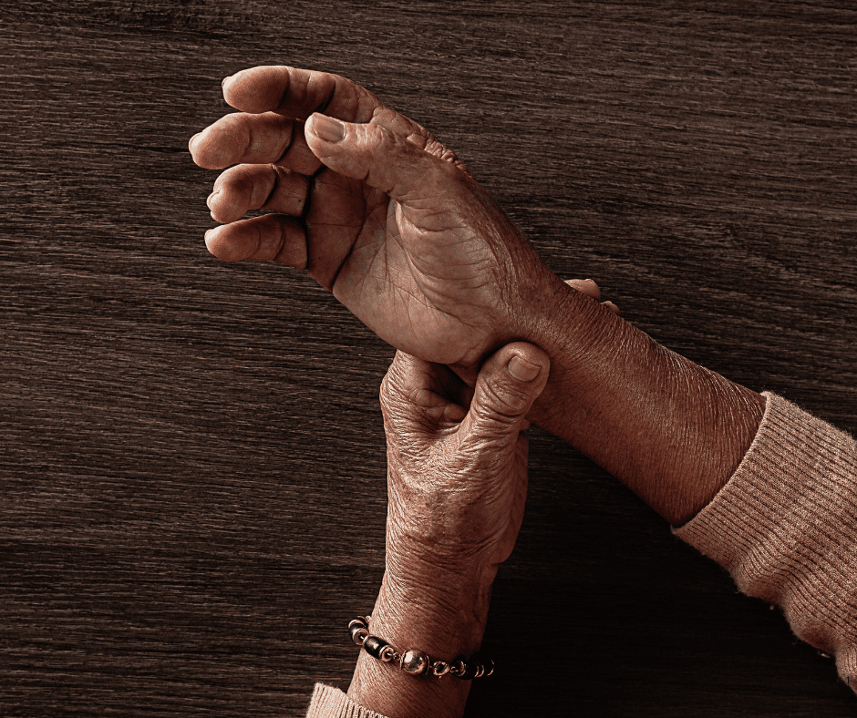 Close-up of an elderly persons hands, one holding the others wrist, against a wooden surface. The person wears a bracelet and a long-sleeve sweater, suggesting care, pain, or tenderness. parkinsons disease
