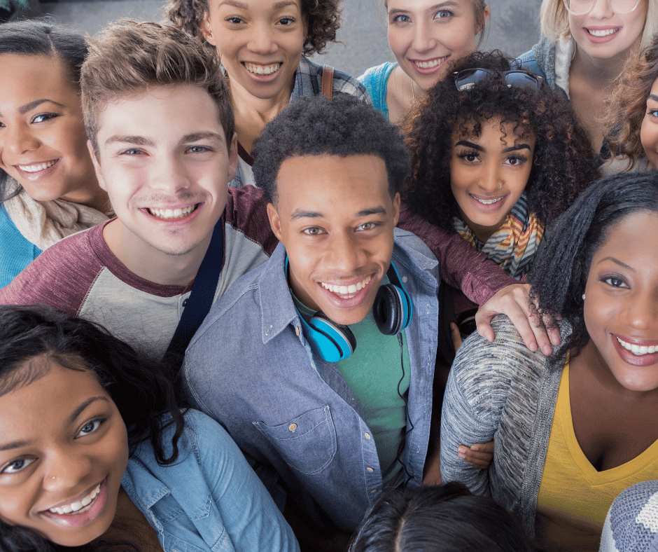 A diverse group of young adults, smiling and standing close together, look up at the camera. One person in front wears blue headphones around his neck. The group appears friendly and cheerful.