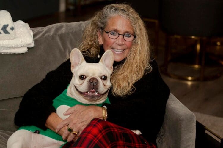 A smiling older woman with long curly hair and glasses sits on a couch, hugging a happy French Bulldog wearing a green sweater. She is dressed in a black sweater and red plaid pants.