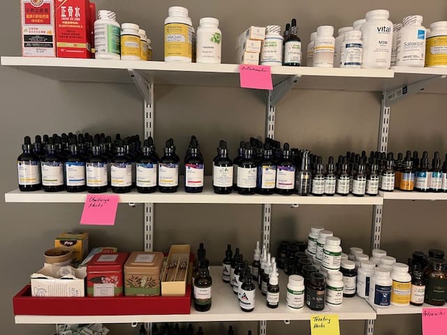 Shelves displaying various health supplements, bottles, and boxes. Several items have handwritten pink and yellow sticky notes labeling categories like antivirals, challenge herbs, and tonify pack.