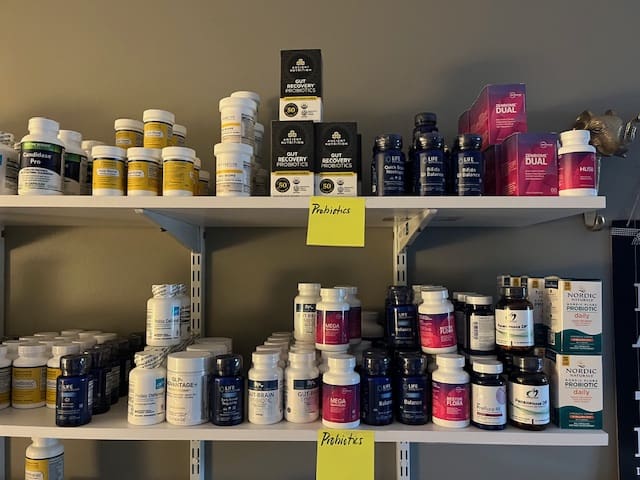 Shelves lined with various supplement bottles and boxes, organized by type. Yellow sticky notes on the middle shelves are labeled Probiotics. The background is a plain gray wall.