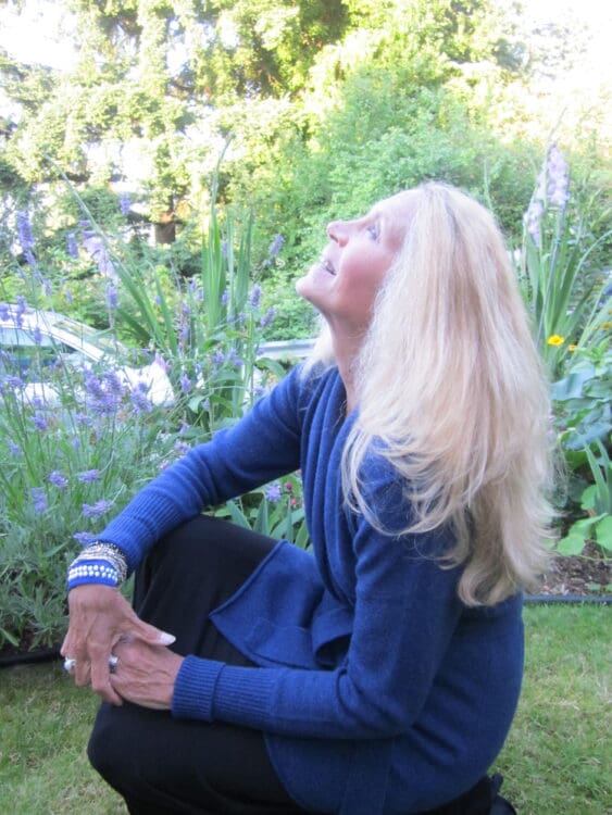 A woman with long blonde hair, wearing a blue sweater and black pants, kneels in a garden surrounded by green plants and purple flowers, looking up and smiling in natural sunlight.