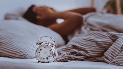 Naturopathic Sleep Doctor: A Natural Approach to Better Rest 2 A classic alarm clock sits on a bed in the foreground, while a person lies under striped sheets in the background, covering their face as if stressed or unable to sleep— naturopathic sleep doctor