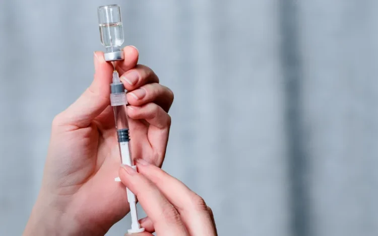 A close-up of hands preparing a syringe by drawing liquid medication from a vial, with a blurred blue-gray background.