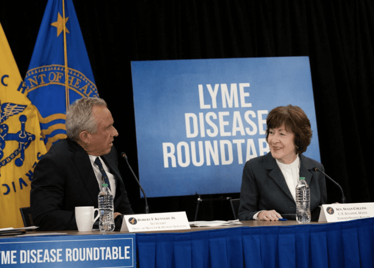Two people sit at a table with microphones, water bottles, and nameplates, speaking at a Lyme Disease Roundtable event. A blue sign and government flags are in the background.
